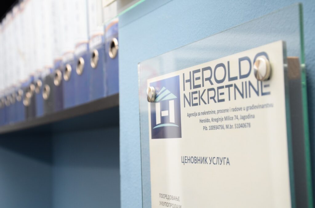 Plaque with "HEROLDO NEKRETNINE" text on glass wall beside a shelf filled with blue binders.